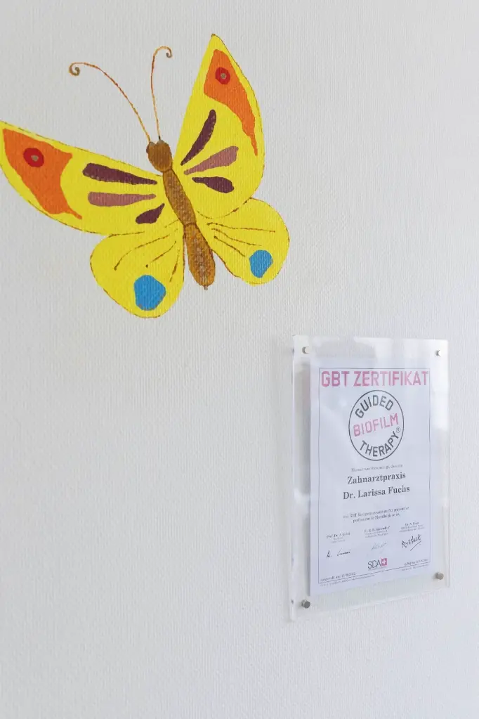 Das GBT-Zertifikat der Zahnarztpraxis Dr. Larissa Fuchs, gerahmt an der Praxiswand neben einem gemalten Schmetterling